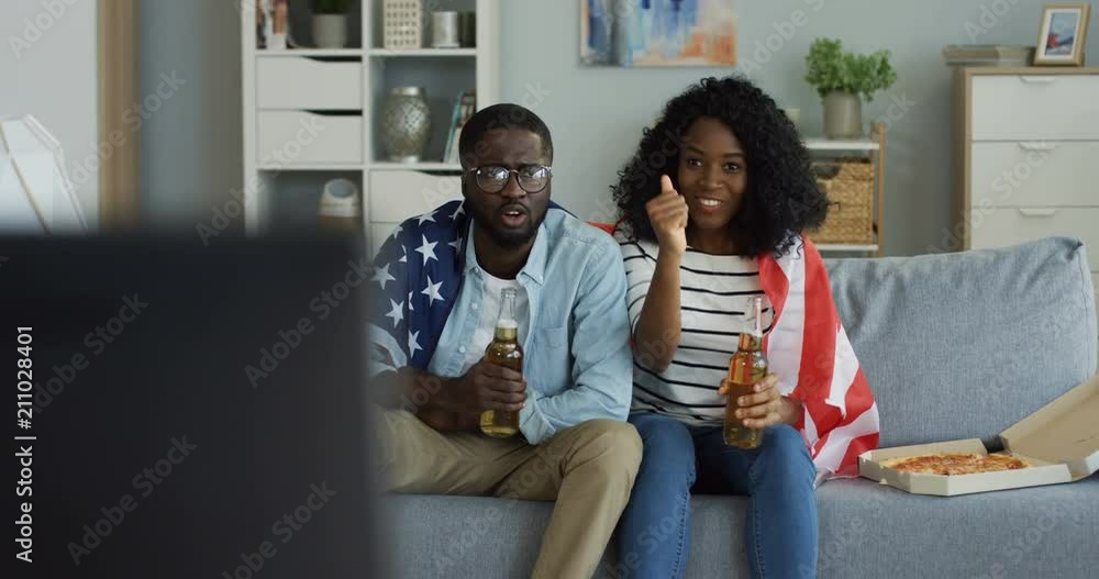 Young African American couple with beer and American flag turning on TV and watching sport channel with positive emotions. At home. Indoor. Young African American couple with beer and American flag turning on TV and watching sport channel with positive emotions. At home. Indoor.