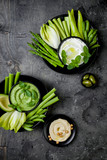 Green vegetables snack board with various dips. Yogurt sauce or labneh, hummus, herb hummus or pesto with fresh vegetables. Healthy raw summer platter.