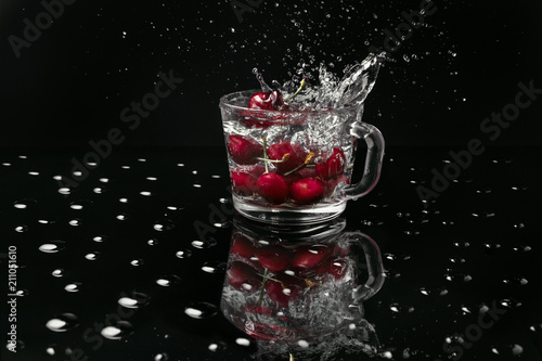 Macro photo of a clear glass mug with cherries in front of a black background on a black table made of glass covered with water drops. 