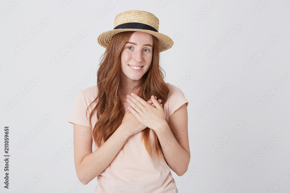 Portrait of happy beautiful young woman in straw hat with long red hair and freckles stands and put hands on her heart isolated over white wall Portrait of happy beautiful young woman in straw hat with long red hair and freckles stands and put hands on her heart isolated over white wall