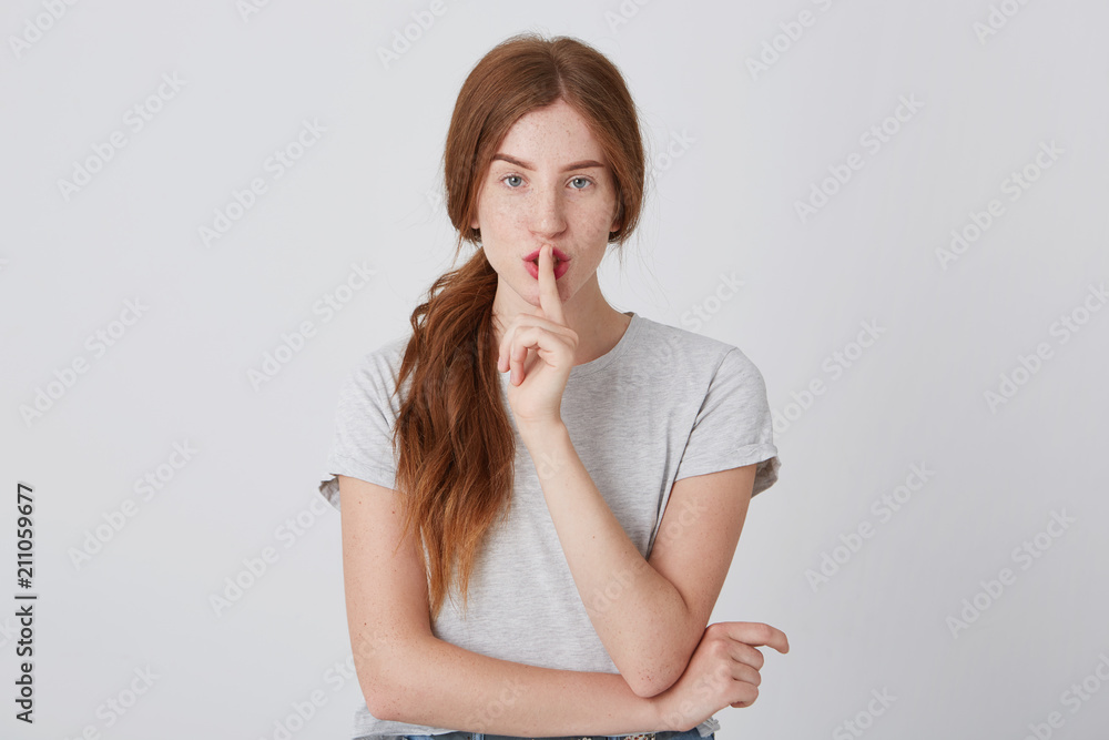 Closeup of attractive alluring young woman with red hair and freckles in tshirt showing silence sign isolated over white background Closeup of attractive alluring young woman with red hair and freckles in tshirt showing silence sign isolated over white background