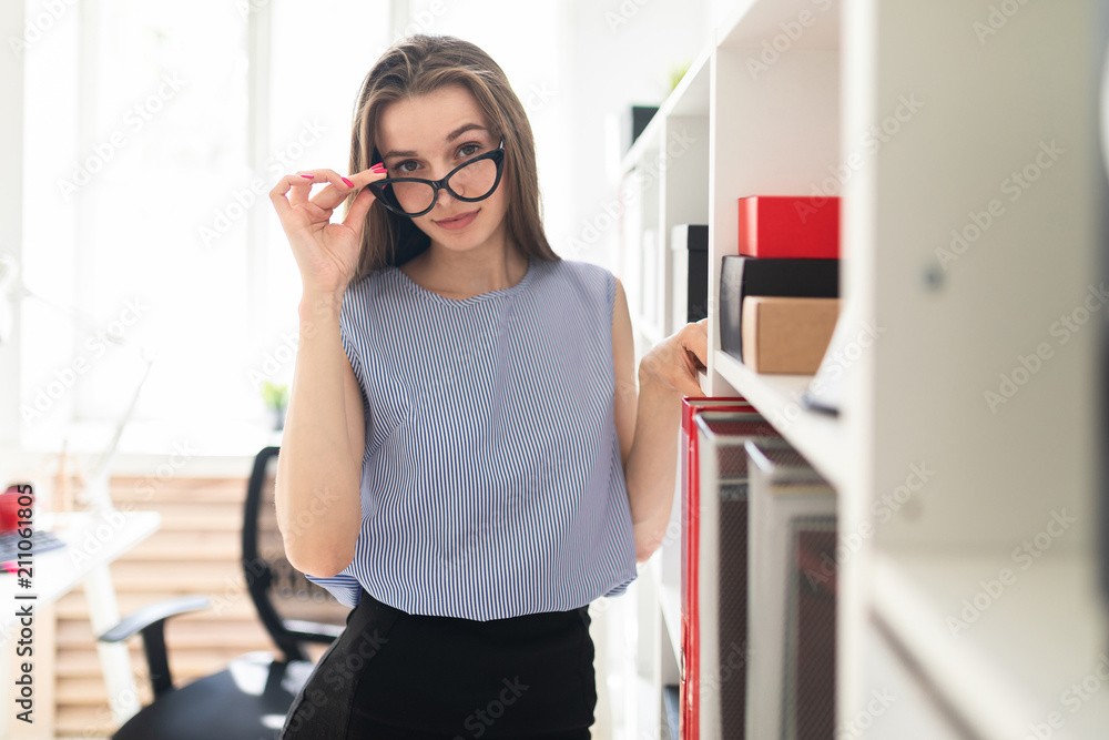 Beautiful young girl in an office stands near a shelf with documents and holds her hand glasses. Beautiful young girl in an office stands near a shelf with documents and holds her hand glasses.