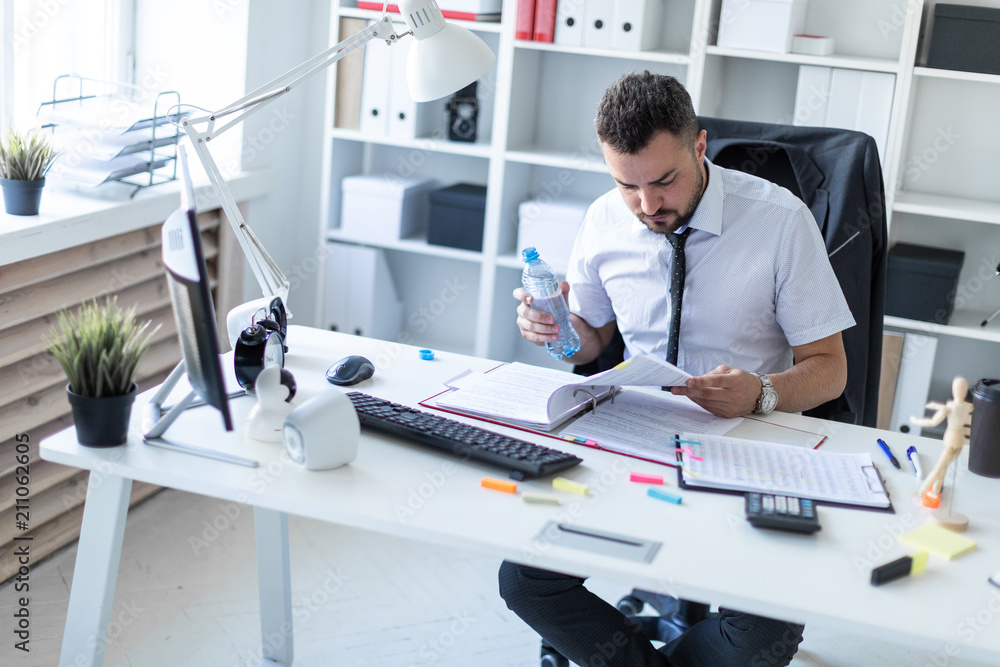 A man is sitting at a table in the office, working with documents and holding a bottle of water in his hand. A man is sitting at a table in the office, working with documents and holding a bottle of water in his hand.