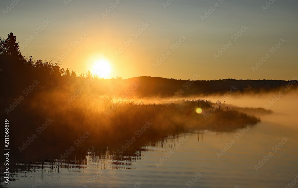Beautiful calm morning foggy sunrise on the Cow Lake near Rocky Mountain House, Alberta, Canada, clear water with mist, reflection of the forest, sun and blue sky. The lake is slowly waking up.