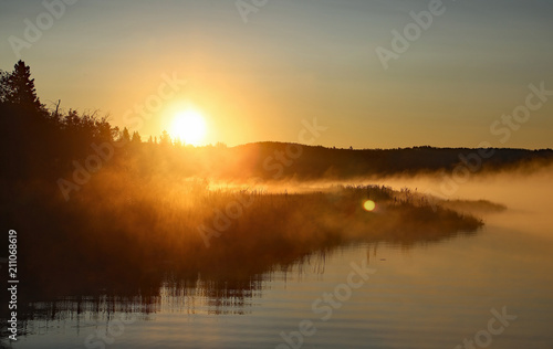 Beautiful calm morning foggy sunrise on the Cow Lake near Rocky Mountain House, Alberta, Canada, clear water with mist, reflection of the forest, sun and blue sky. The lake is slowly waking up. © Jitka