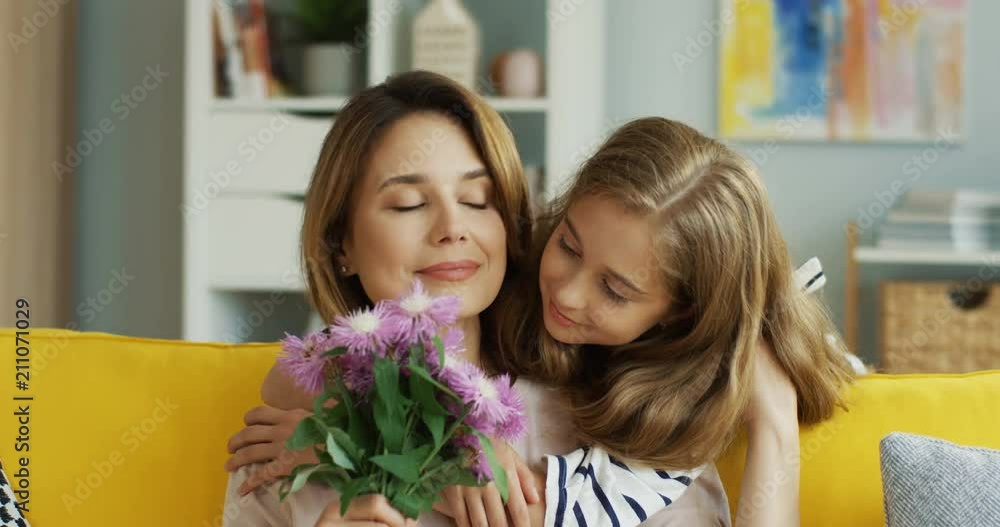 Close up of the beautiful caucasian mother and daughter on the sofa hugging and woman holding a bouquet of flowers her daughter gave her. Portrait. Inside. Close up of the beautiful caucasian mother and daughter on the sofa hugging and woman holding a bouquet of flowers her daughter gave her. Portrait. Inside.