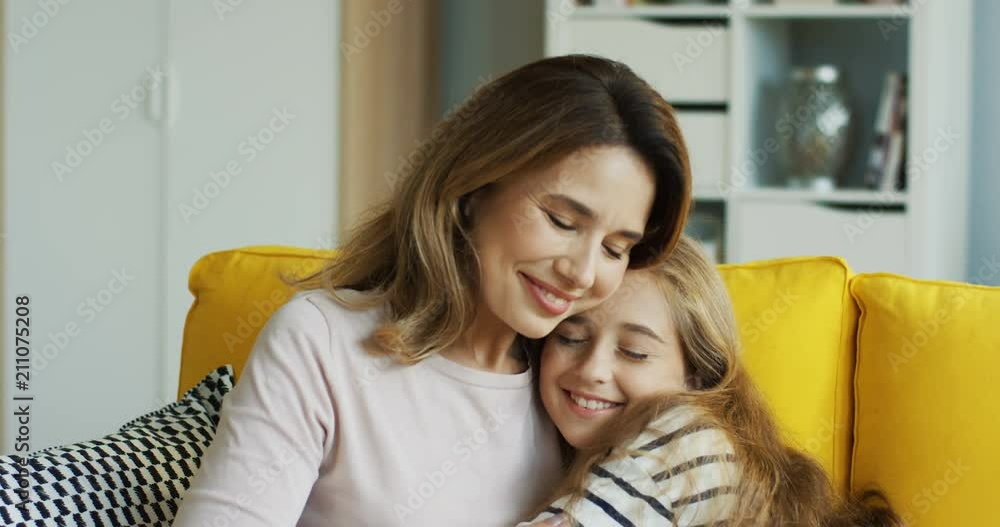 Close up of the teenage pretty girl coming to her mother who sitting on the yellow sofa in the cozy room and embracing her. At home. Inside. Close up of the teenage pretty girl coming to her mother who sitting on the yellow sofa in the cozy room and embracing her. At home. Inside.