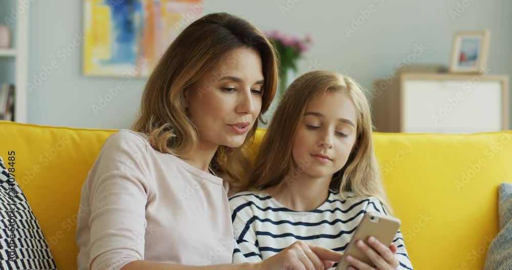 Portrait of the Caucasian pretty little girl with her mother scrolling, taping on the smartphone and talking while sitting in the living room. Indoors. Portrait of the Caucasian pretty little girl with her mother scrolling, taping on the smartphone and talking while sitting in the living room. Indoors.
