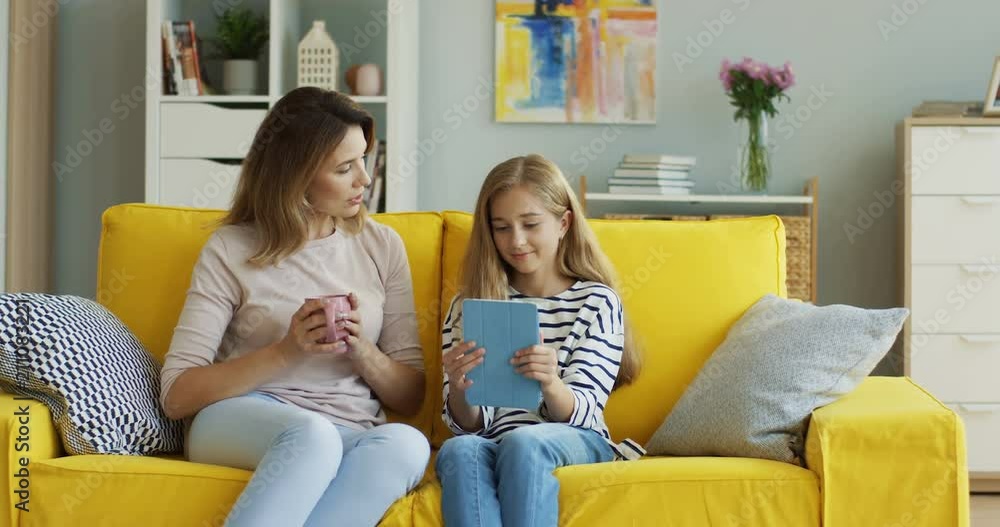 Caucasian beautiful woman drinking tea or coffee and talking with her teenage daughter who using a tablet computer in the living room. Indoor. Caucasian beautiful woman drinking tea or coffee and talking with her teenage daughter who using a tablet computer in the living room. Indoor.