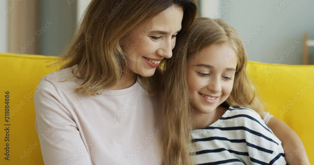 Close up of the caucasian cheerful mother and daughter watching video on the tablet device while resting on the sofa in the modern living room. Indoor. Close up of the caucasian cheerful mother and daughter watching video on the tablet device while resting on the sofa in the modern living room. Indoor.