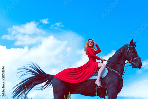 Beautiful young woman in long red dress riding a horse in countryside. Portrait of a dark horse and woman. Attractive girl riding on horse rural location 