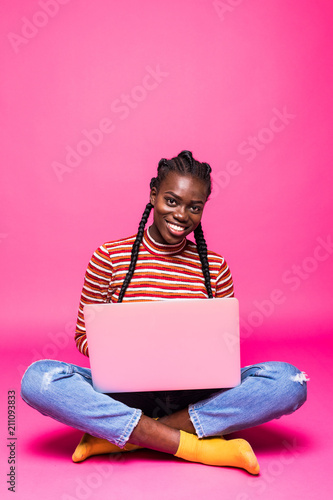 Attractive afro american woman with beautiful smile using silver notebook, while sitting in lotus pose on the floor isolated over pink background photo