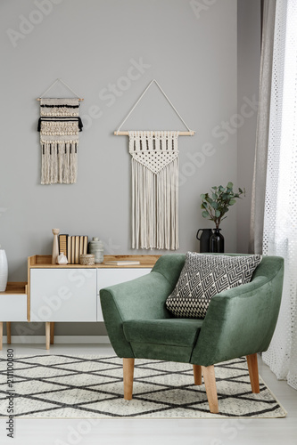 Real photo of a boho living room interior with macrame hanging on gray wall behind a comfy, green armchair with a cushion