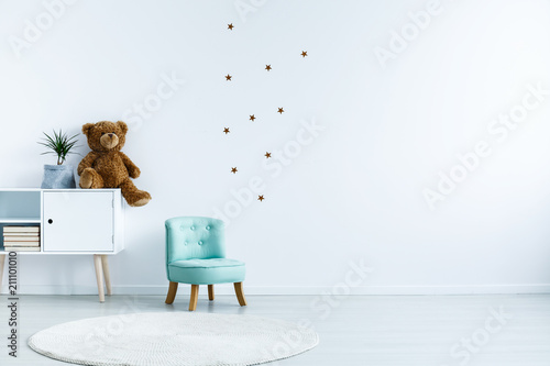 Small light blue armchair for kid standing in white room interior with stars on the wall, white rug and cupboard with books, teddy bear and fresh plant. Empty space for your crib