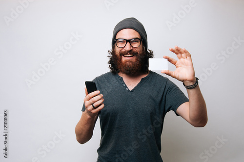 Cheerful smiling bearded hipster man holding smartphone and credit card, online shopping concept photo