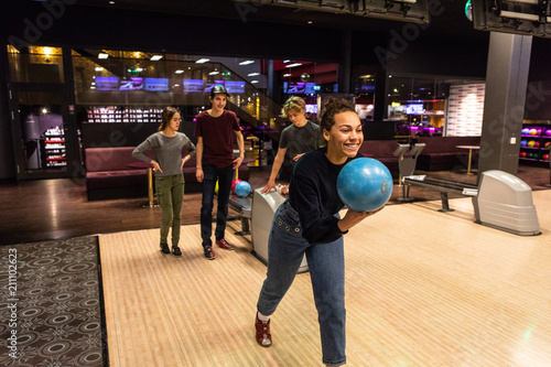 Smiling teenage girl holding ball against friends standing on illuminated parquet floor at bowling alley photo