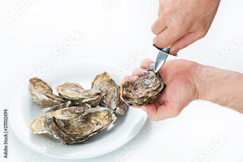 Fresh oyster. Man open fresh oyster. Raw fresh oyster is on white round plate, image isolated, with soft focus. Restaurant delicacy. Saltwater oyster.