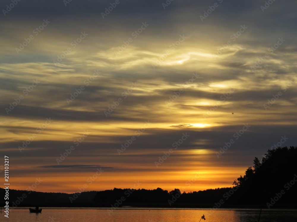 Fisherman on Boat Behind Reeds at Sunset Over Beautiful Lake with Cloudy Sky in background Fisherman on Boat Behind Reeds at Sunset Over Beautiful Lake with Cloudy Sky in background