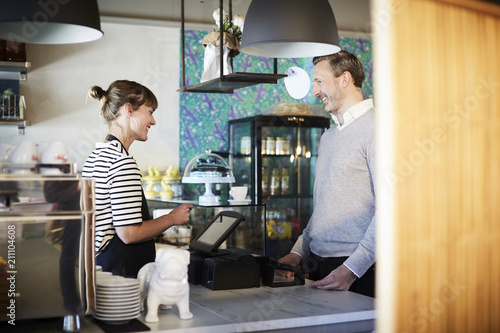 Smiling barista talking to customer while using cash register at checkout counter in cafe