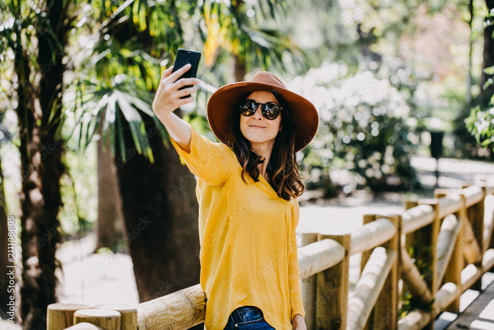 Obraz premium .Pretty young woman wearing a brown hat walking around a famous park in Madrid in a summer day using her mobile phone, talking and texting. Lifestyle portrait.