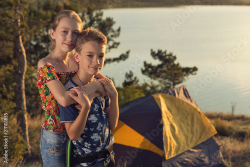 two children, a boy and a girl are standing near a tent on the background of a lake or a bay, a girl is standing behind and hugging a boy, a bivouac or camping, a summer trip, summer, family
