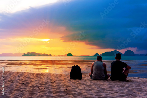 Back of young caucasian men sitting on the beach watching sunrise. Couple gay enjoying beautiful sunset with copy space. Two American people on summer holidays in Tup Kaek Beach, Krabi, Thailand. photo