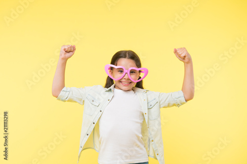Loves power. Kid girl heart shaped eyeglasses cheerful posing with power gesture. Girl curly hairstyle adorable smiling face. Child charming smile yellow background. Kid happy lovely enjoy childhood