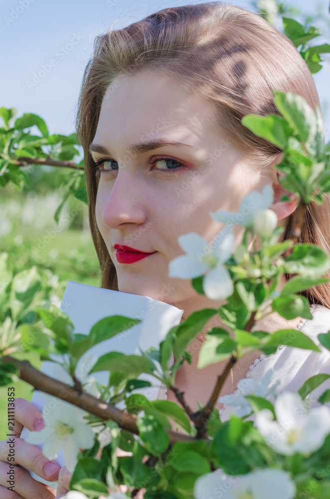 Close-up young pretty woman in white top standing under blossoming apple tree and holds a book on sunny day. Close-up young pretty woman in white top standing under blossoming apple tree and holds a book on sunny day.