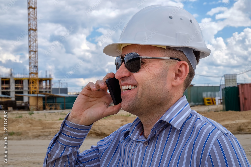 The foreman in white helmet on the construction site speaks on a mobile phone and smiles. The concept of a successful businessman The foreman in white helmet on the construction site speaks on a mobile phone and smiles. The concept of a successful businessman