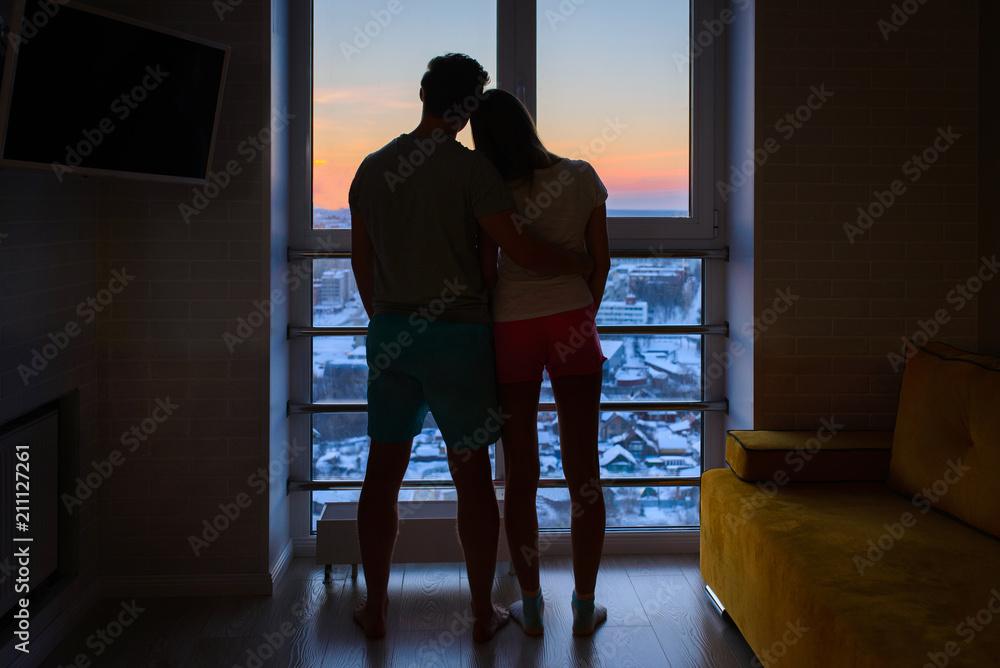 Yong couple in love standing close to window during the sunrise, silhouette full length shoot.