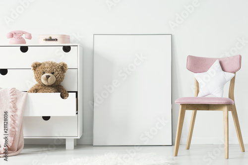 Empty poster with place for your graphic standing on the floor in white room interior with pink wooden chair and teddy bear placed in cupboard's shelf