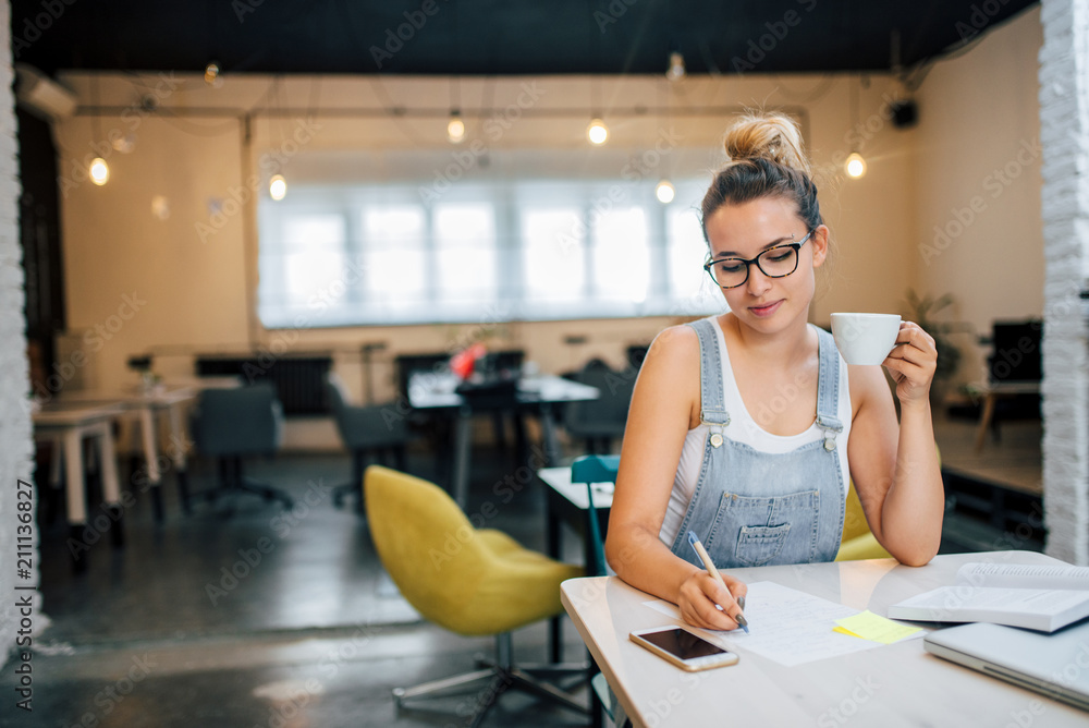 Young woman writing on paper while holding a cup. Young woman writing on paper while holding a cup.