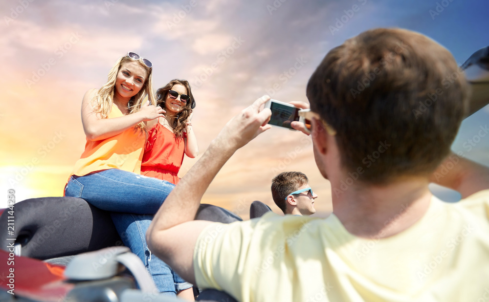 Fototapeta premium leisure, road trip, travel and people concept - happy friends taking picture by smartphone in convertible car over sky background