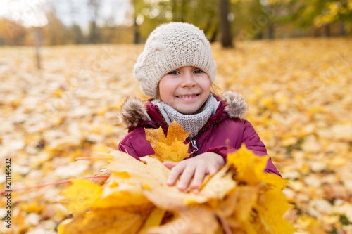 childhood  season and people concept - happy little girl with fallen maple leaves at autumn park