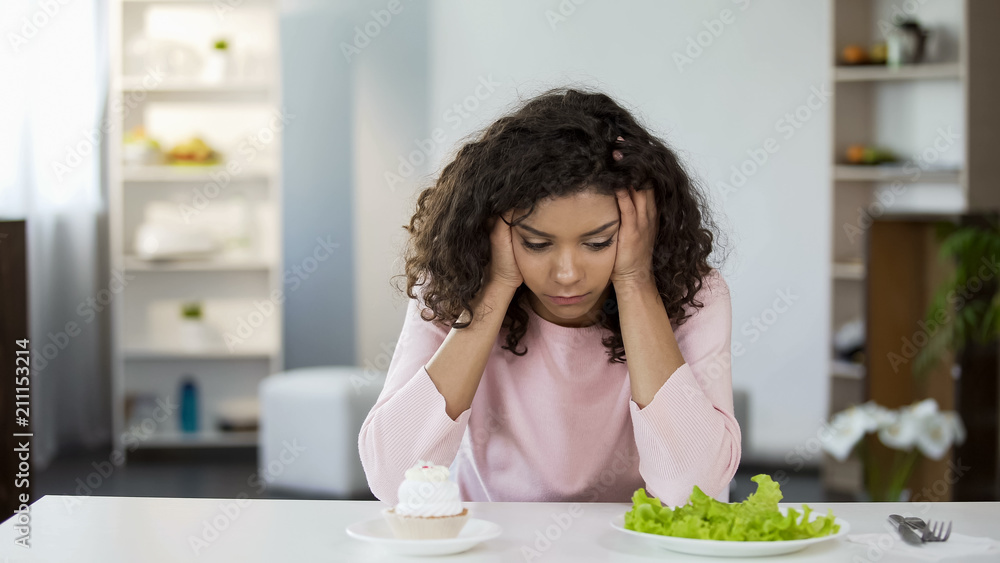 Young biracial woman choosing between salad and sweets, health care choice