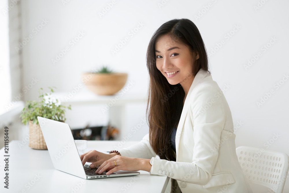 Portrait of beautiful Asian female office worker looking at camera smiling while working at laptop, young businesswoman posing for company catalogue near desk. Concept of leadership, ethnicity Portrait of beautiful Asian female office worker looking at camera smiling while working at laptop, young businesswoman posing for company catalogue near desk. Concept of leadership, ethnicity