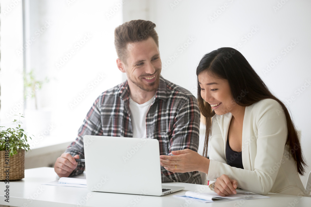 Smiling diverse colleagues laughing at reading funny news at laptop while working together at shared space, employees excited by unexpected business success or rates growth. Cooperation concept