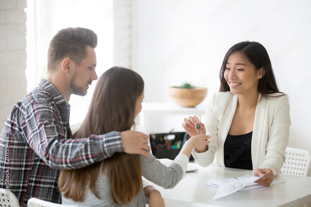 Smiling Asian broker giving keys to millennial couple, buying first home together, female real estate agent presenting keys to young spouses deciding to purchase property. New beginning concept Smiling Asian broker giving keys to millennial couple, buying first home together, female real estate agent presenting keys to young spouses deciding to purchase property. New beginning concept