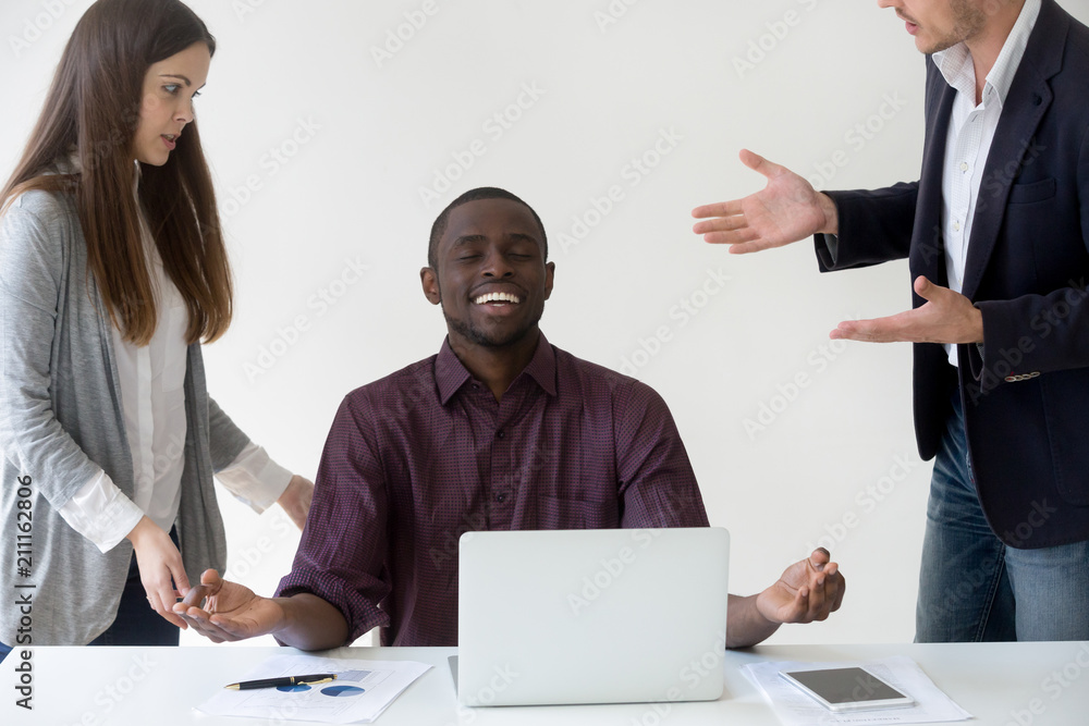 Happy African American employee meditating at desk, reaching nirvana state ignoring colleagues talking to him, black worker abstracting from annoying colleagues meditating. Stress relief concept Happy African American employee meditating at desk, reaching nirvana state ignoring colleagues talking to him, black worker abstracting from annoying colleagues meditating. Stress relief concept