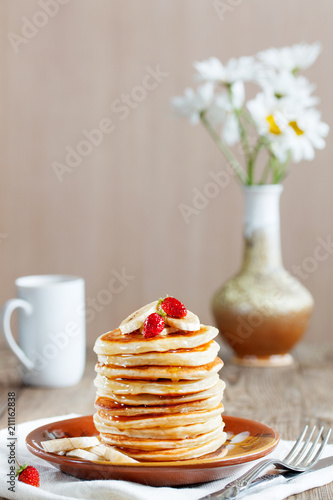 Pancakes with banana slices, strawberries sprinkled with honey for a breakfast. Camomiles and white mug in the background.