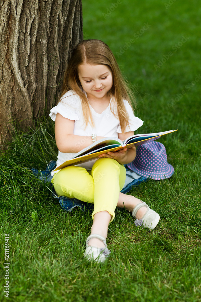 Little girl reading a book sitting on a grass under a tree summer day outdoors.

