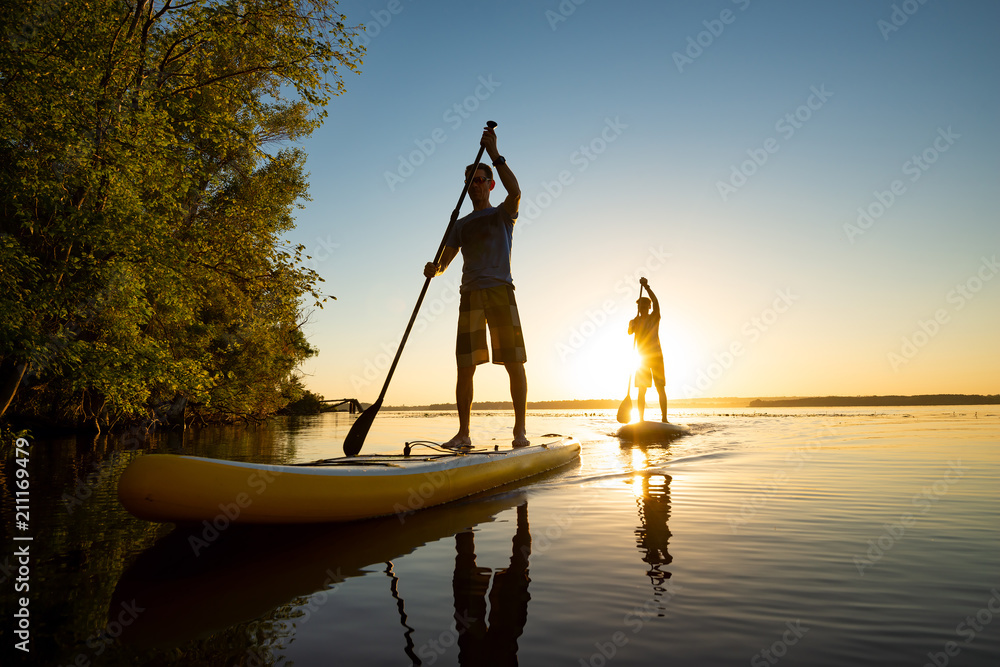 Men, friends sail on a SUP boards in a rays of rising sun