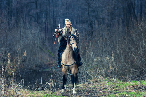 Scandinavian northern viking riding horse with ax in hand. Northern warrior woman riding in forest in war clothes with fur collar, war makeup.