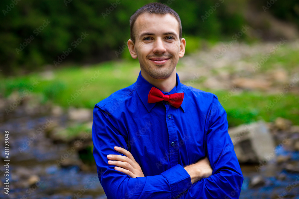 Portrait of young smiling handsome man in blue shirt near forest river. Emotions concept Portrait of young smiling handsome man in blue shirt near forest river. Emotions concept