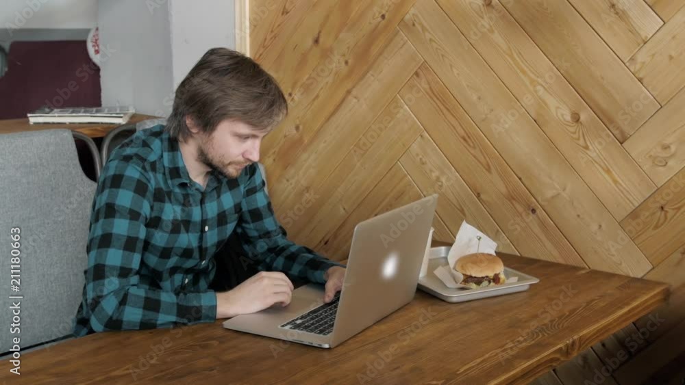 Portrait of busy young business man sitting with glass of drink and hamburger in fast food restaurant interior. Attractive caucasian eating and using notebook computer in cafe. Portrait of busy young business man sitting with glass of drink and hamburger in fast food restaurant interior. Attractive caucasian eating and using notebook computer in cafe.