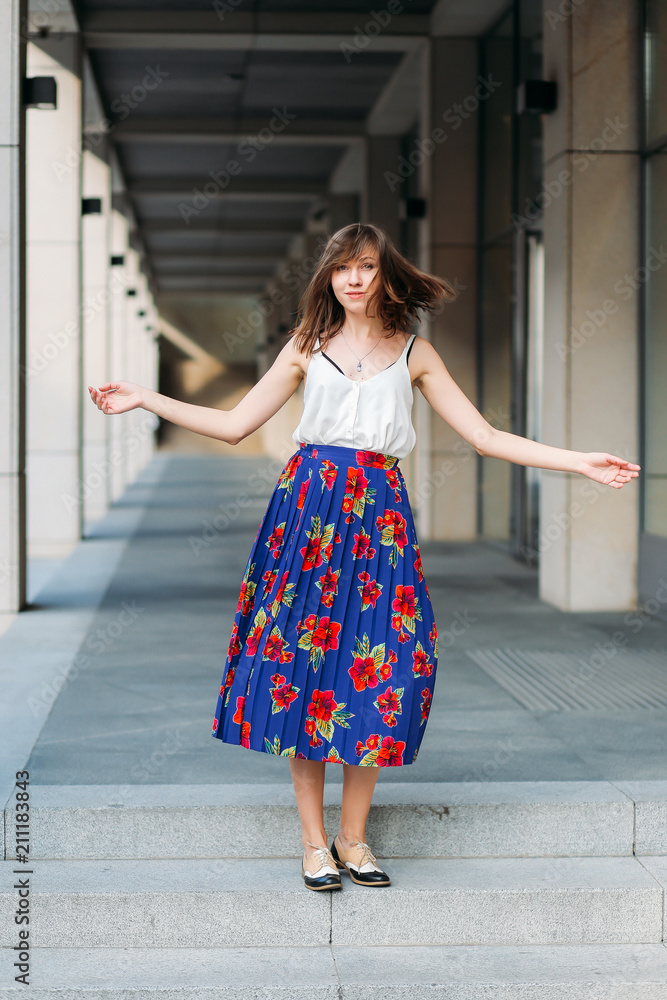 Stylish woman dancing on the street. Fashion portrait of beautiful woman on the street, urban background. Woman portrait outdoors in floral skirt and white top.