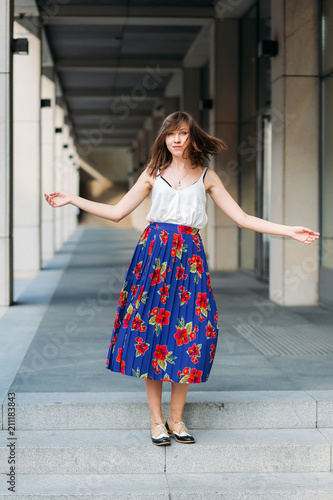 Stylish woman dancing on the street. Fashion portrait of beautiful woman on the street, urban background. Woman portrait outdoors in floral skirt and white top. © Marharyta