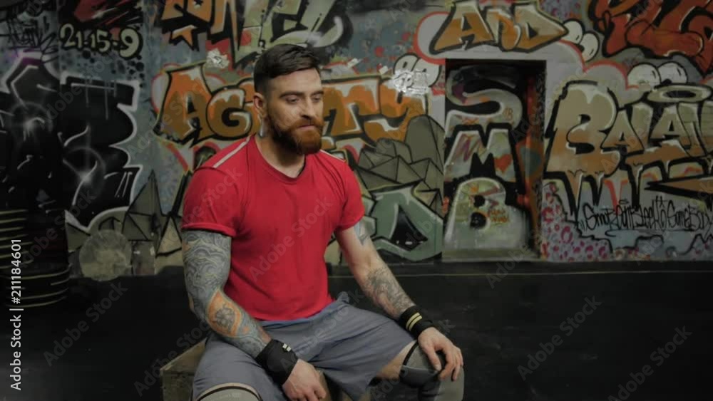 Muscular man with a beard raises a barbell above his head in the split squat in the gym on graffiti wall background. Handsome male athlete professional