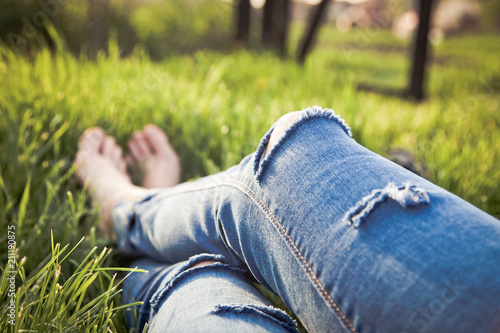 legs of hipster woman wearing torn jeans lying in green grass with smartphone and headsets enjoying sunset - summertime relaxation