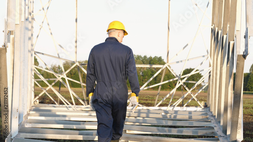 An electrician or builder, an employee in a blue robe, wearing a yellow helmet, is preparing to install electric poles, an electric tower, and energy.
